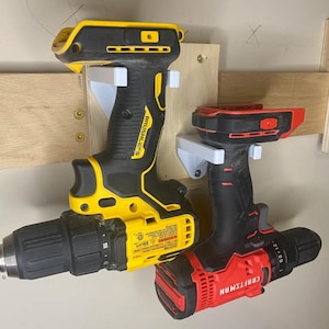 May include: Two cordless drills mounted on a wall-mounted rack. The drill on the left is yellow and black with the word "BRUSHLESS" printed on the side. The drill on the right is red and black with the word "CRAFTSMAN" printed on the side.
