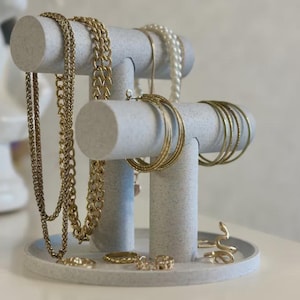 May include: A jewellery display stand made of speckled grey material. Gold necklaces, bracelets, and rings are displayed on the stand. A pearl necklace and gold hoop earrings are also visible. The stand has a round base and two horizontal bars.