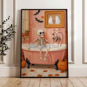 K&ouml;nnte beinhalten: Ein gerahmter Halloween-Druck, der ein Skelett in einer rosa Badewanne mit Blasen zeigt. Das Kunstwerk umfasst Flederm&auml;use, K&uuml;rbisse und ein Gem&auml;lde von zwei Geistern, wodurch eine gruselige und doch skurrile Atmosph&auml;re entsteht. Der Druck ist vor einem rosa Hintergrund.