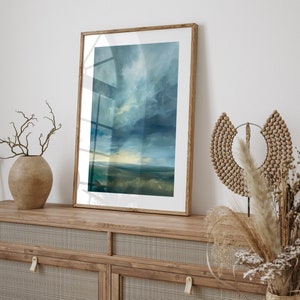 May include: A framed landscape art print featuring a moody sky and field in shades of blue, green, and yellow. The artwork is displayed on a wooden cabinet with a woven cane front, alongside decorative vases and dried plants.