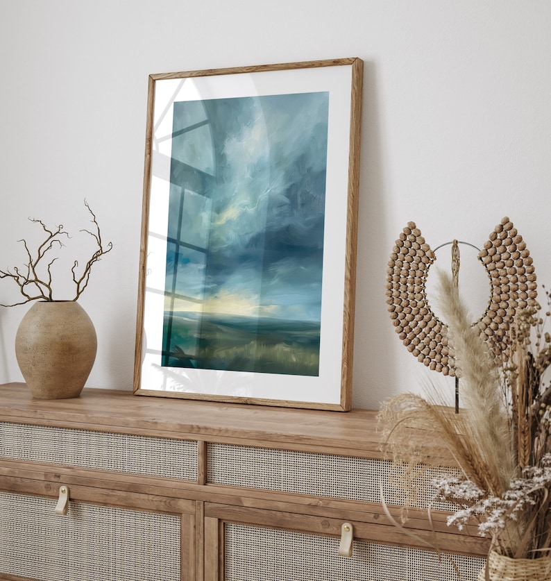 May include: A framed landscape art print featuring a moody sky and field in shades of blue, green, and yellow. The artwork is displayed on a wooden cabinet with a woven cane front, alongside decorative vases and dried plants.