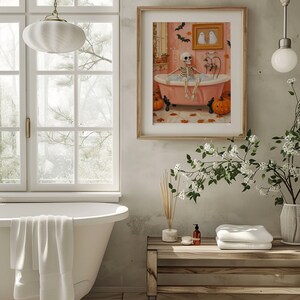 K&ouml;nnte beinhalten: Ein gerahmter Druck zeigt ein Skelett, das in einer rosa Badewanne mit Blasen entspannt. Die Badezimmerszene umfasst K&uuml;rbisse, Flederm&auml;use und ein gerahmtes Bild von zwei Geistern, was ein Halloween-Thema hervorruft. Das Kunstwerk ist in einem Badezimmer mit einer wei&szlig;en Badewanne.
