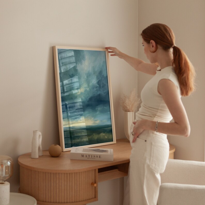May include: A framed landscape painting with a light wood frame. The artwork features a moody sky with shades of blue and green over a field. The painting is displayed on a wooden desk.