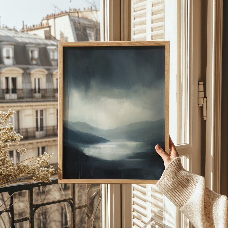 May include: A framed painting of a landscape with a blue and gray color scheme. The painting depicts a misty, mountainous scene with a lake in the foreground. The frame is made of wood and has a light brown finish.