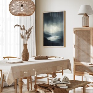 May include: A light brown wicker pendant lamp hangs over a dining table with a beige tablecloth. The table is set with a wooden bowl and a white plate. A wicker vase with dried flowers sits on the table. A framed abstract painting of a blue landscape hangs on the wall behind the table. A wicker cabinet with a wooden top sits to the right of the table. A brown throw blanket is draped over a wicker bench with a stack of books on top.