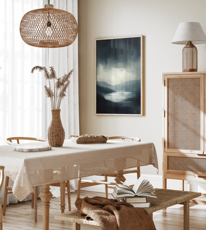 May include: A light brown wicker pendant lamp hangs over a dining table with a beige tablecloth. The table is set with a wooden bowl and a white plate. A wicker vase with dried flowers sits on the table. A framed abstract painting of a blue landscape hangs on the wall behind the table. A wicker cabinet with a wooden top sits to the right of the table. A brown throw blanket is draped over a wicker bench with a stack of books on top.