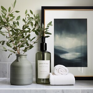 May include: A bathroom vanity with a green vase filled with eucalyptus branches, a clear glass bottle with a black pump labeled "Olive & Oak", and a white rolled towel. A framed print of a blue and gray landscape hangs on the wall behind the items.
