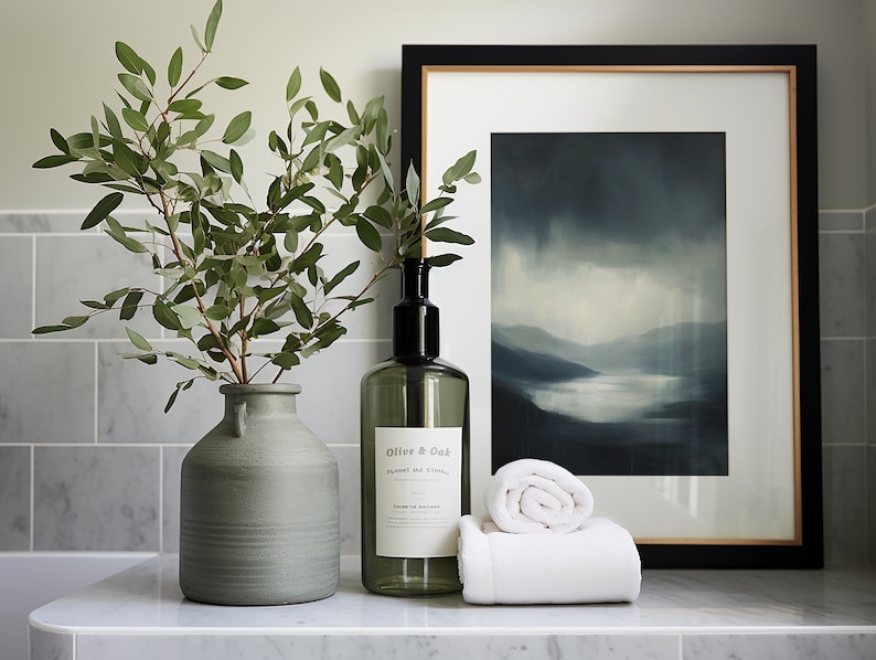 May include: A bathroom vanity with a green vase filled with eucalyptus branches, a clear glass bottle with a black pump labeled "Olive & Oak", and a white rolled towel. A framed print of a blue and gray landscape hangs on the wall behind the items.
