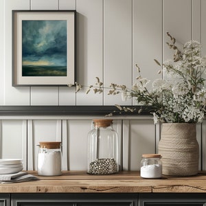 May include: A framed landscape painting with a stormy sky hangs above a wooden countertop. Clear glass jars with cork lids contain ingredients. A woven vase holds white flowers, and a stack of white plates sits nearby.
