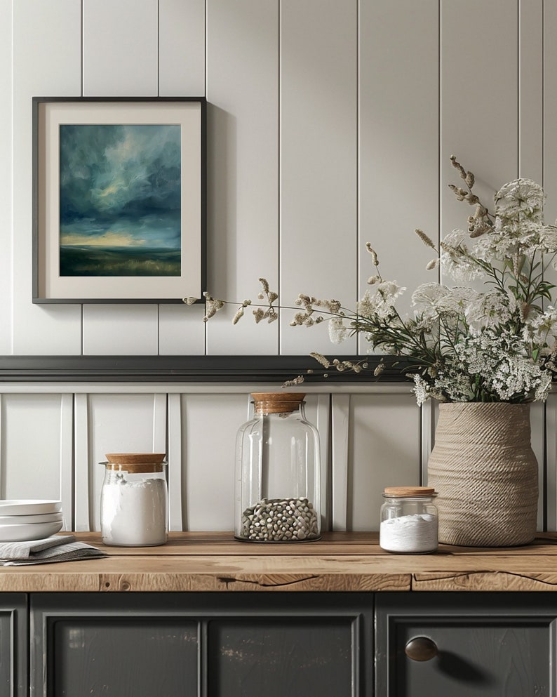 May include: A framed landscape painting with a stormy sky hangs above a wooden countertop. Clear glass jars with cork lids contain ingredients. A woven vase holds white flowers, and a stack of white plates sits nearby.