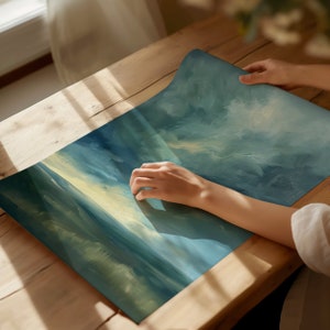 May include: A rolled-up, rectangular art print with a blue and yellow abstract landscape design. The print is being held on a wooden table. The artwork features brushstrokes and a gradient of colors, evoking a sense of depth and atmosphere.