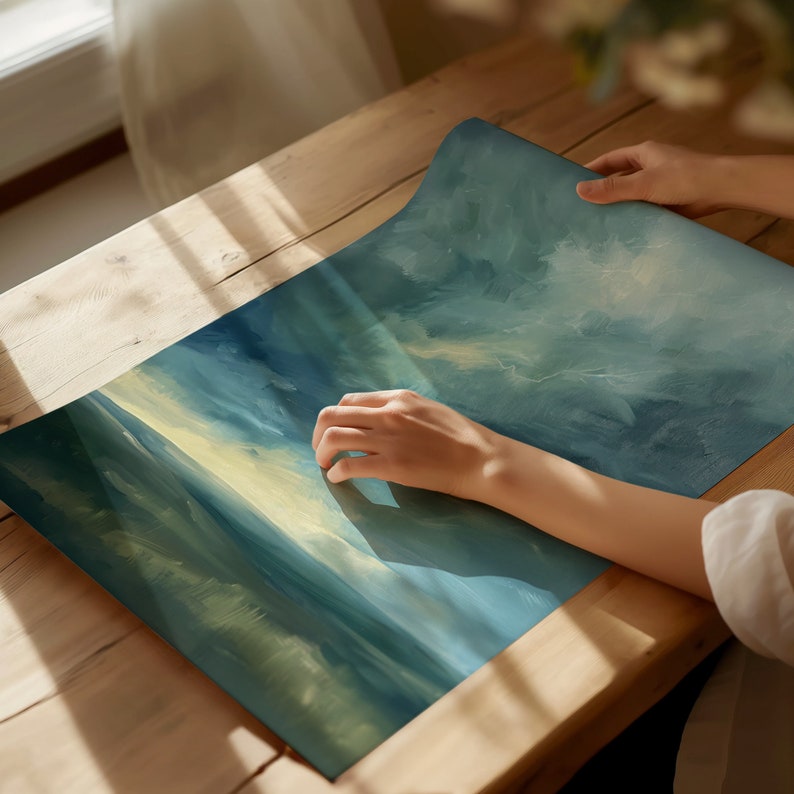 May include: A rolled-up, rectangular art print with a blue and yellow abstract landscape design. The print is being held on a wooden table. The artwork features brushstrokes and a gradient of colors, evoking a sense of depth and atmosphere.