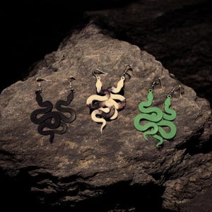 May include: Three pairs of snake-shaped earrings in black, cream and black, and green. The earrings are displayed on a dark grey rock surface. Each pair has a different colour and design, with a hook for pierced ears.