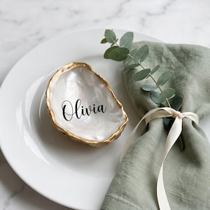 Puede incluir: Un plato de cerámica blanca con una concha de ostra nacarada con borde dorado y el nombre Olivia impreso en letra cursiva negra. Junto a la concha hay una servilleta de lino verde salvia atada con una cinta color crema y una rama de eucalipto.