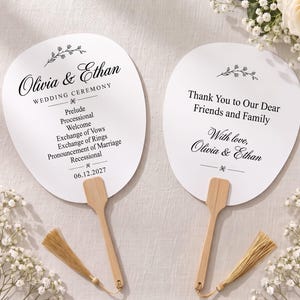 Op de afbeelding: Twee witte waaiers in peddelvorm voor een huwelijksceremonie met houten handvatten en lichtbruine kwastjes. De linkerwaaier toont de tekst Olivia & Ethan, Wedding Ceremony, een programma en de datum 06.12.2027. De rechterwaaier vermeldt Thank You to Our Dear Friends and Family, With love, Olivia & Ethan.