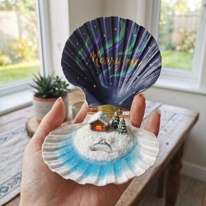 Könnte beinhalten: Eine dekorative Muschel-Ringbox mit handbemaltem dunkelblauem Innenraum, grünen und violetten Nordlichtmustern und dem goldenen Schriftzug Marry me. Die Basis enthält eine winterliche Miniaturlandschaft mit einer Hütte und zwei Tannen.