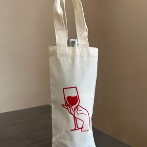 Op de afbeelding: Een crèmekleurige canvas wijnfleszak met een rode lijntekening van een hand die een wijnglas vasthoudt. De hoge, rechthoekige stoffen drager heeft twee bijpassende handvatten en bevat een enkele fles met een zilverkleurige metalen dop.
