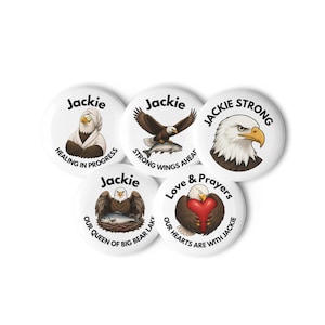 Op de afbeelding: Vijf ronde witte buttons met illustraties van Amerikaanse zeearenden en tekst. De ontwerpen tonen Jackie met een handdoek, vliegend met een vis, een portret met Jackie Strong, zittend in een nest met een vis, en met een rood hart en Love & Prayers.