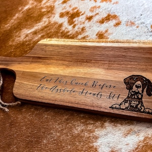 May include: Wooden cutting board with a handle and rope detail. The board features engraved text that reads "Eat This Quick Before The Asshole Steals It!" and a dog graphic. The board is brown.
