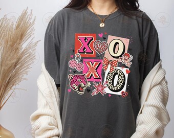 May include: A charcoal gray short-sleeve t-shirt featuring a graphic print with the letters X and O, a disco ball heart, pink dice, a red polka dot bow, a cupid figure, a leopard print letter O, and a pink leopard head with star accents.