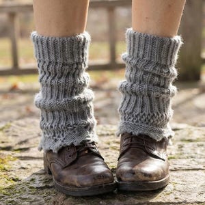 Puede incluir: Un par de calentadores de punto gris con textura acanalada y borde festoneado, usados sobre botas de cuero marrón oscuro con cordones. Los calentadores presentan un patrón de punto vertical que se extiende desde el tobillo hasta la pantorrilla.