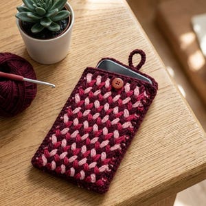 May include: A rectangular crochet phone pouch featuring a woven pattern in shades of burgundy and light pink. The pouch includes a small brown button closure at the top centre and a matching crochet loop for hanging or carrying the accessory.