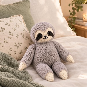 May include: A plush sloth toy crafted from textured grey yarn with cream-coloured hands and feet. The sloth features a round head, dark circular eyes, and a stitched black nose and mouth on a cream-coloured face patch with dark eye markings.
