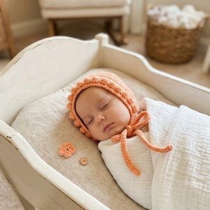 May include: A peach-colored crocheted baby bonnet with a textured bobble trim and tie strings, paired with a small crocheted flower and a wooden button. The bonnet is worn by a sleeping infant wrapped in a white muslin swaddle blanket.