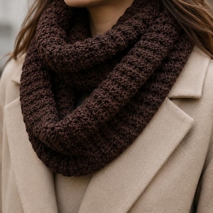May include: A dark brown chunky knit infinity scarf with a textured, ribbed pattern worn around the neck over a beige wool coat. The scarf features a thick, looped design that sits close to the collar, showcasing a consistent, dense stitch.