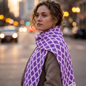 Pode incluir: Um cachecol longo de malha com um padrão repetitivo de diamantes em roxo e branco. O cachecol é finalizado com franjas brancas em ambas as extremidades. O tecido tem uma aparência texturizada e o design é consistente em todo o comprimento.