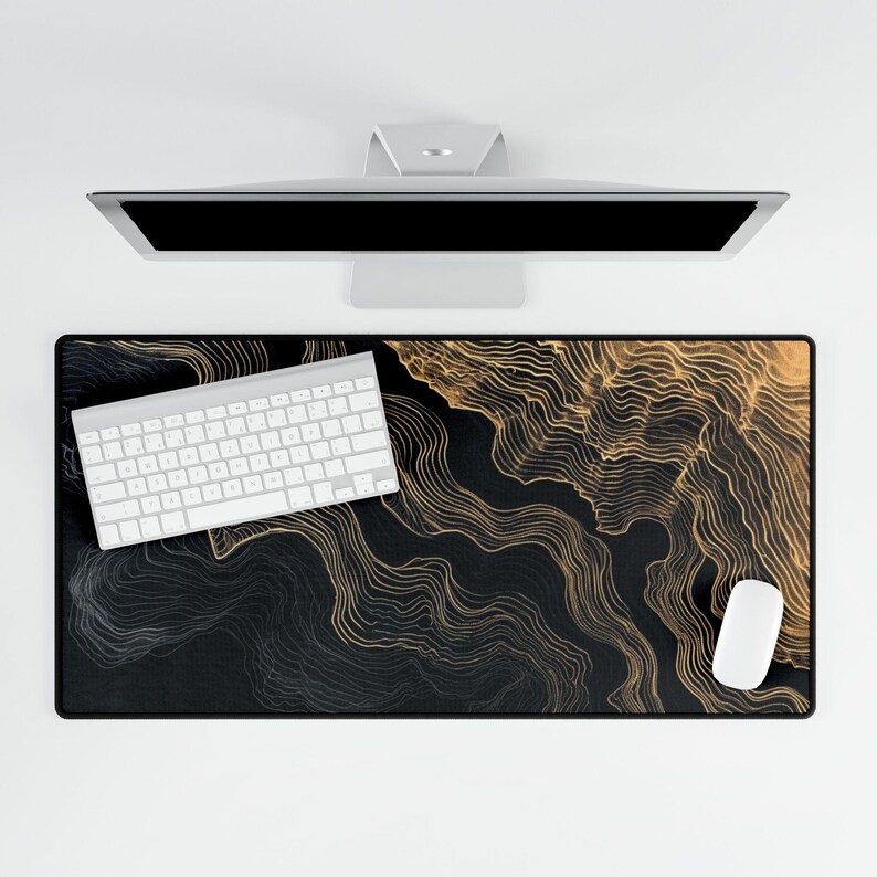 A black and gold patterned desk mat with a white keyboard and mouse. The mat features a flowing, abstract design in gold against a black background. A computer monitor is visible above the mat.