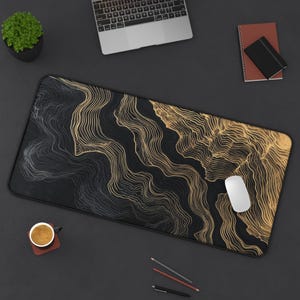 A large black and gold abstract design desk mat. The rectangular mousepad features a flowing, topographic-like pattern in gold against a black background. A white mouse is on the right side of the mousepad.