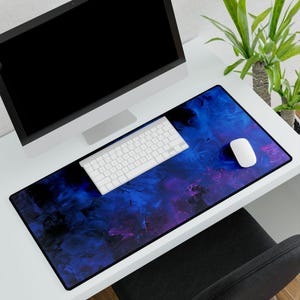 May include: A rectangular desk mat with a vibrant abstract design in shades of blue, purple, and black. A white keyboard and mouse sit on the mat, alongside a computer monitor. The mat adds a decorative touch to a workspace.