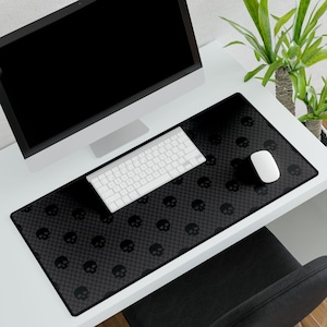 May include: A black desk mat with a repeating skull pattern, a white keyboard, and a white mouse. The mat is rectangular and covers a portion of a white desk. A computer monitor is also on the desk.