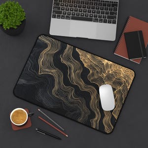 A black and gold abstract design mousepad with a black border. A white computer mouse sits on the mousepad. A laptop, notebooks, pencils, and a cup of coffee are also on the dark surface.