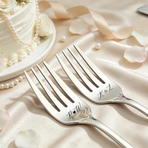 May include: Two silver forks with engraved initials, set on a satin surface, with a wedding cake and rose petals in the background. The cake is white with pearl accents and roses. One fork has "K & Z" and the other has "R & W" engraved.