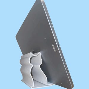 May include: A gray tablet computer is propped up on a white cat-shaped stand. The stand is made of a textured material and has a cutout in the shape of a cat.
