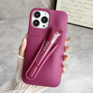 May include: A magenta phone case with the brand name "rhode" embossed on the side. A matching magenta lip balm tube with the word "mode" is attached. The lip balm is labeled "Peptide Lip Tint".