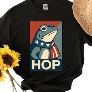 Puede incluir: Camiseta negra con un gráfico de rana y la palabra "HOP". La rana es azul y lleva una bufanda con estrellas y rayas. Un sombrero de paja y girasoles están en el fondo.