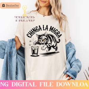 May include: Cream-colored t-shirt featuring a black and white illustration of a cat swatting at a drink with ice cubes. The text "CHINGA LA MIGRA" is above the cat. The shirt also has a "Bestseller" and "Trend Winners Wear" logo.