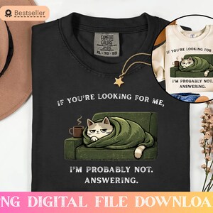 Puede incluir: Camiseta negra con el texto "If you're looking for me, I'm probably not answering". El diseño muestra un gato gruñón envuelto en una manta en un sofá verde, con una taza de café. También se ve un collar con un colgante de estrella dorada.