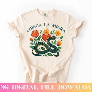 May include: Cream-colored t-shirt featuring a graphic design with the text "CHINGA LA MIGRA". The design includes a green snake, a red heart, orange flowers, and green leaves. The shirt is made of a soft material.