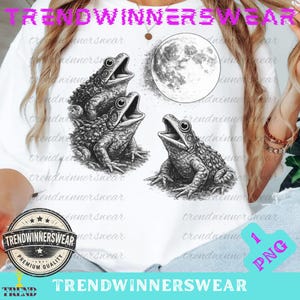 May include: White t-shirt with a black and white illustration of three frogs and a full moon. The design has a detailed, artistic style, suitable for those who appreciate nature and vintage aesthetics.