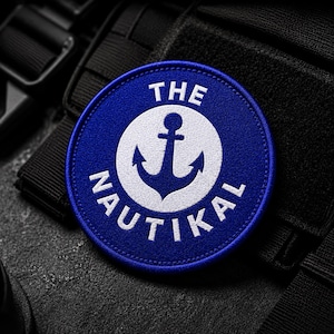 May include: A circular blue patch featuring a white anchor graphic in the center. The text THE NAUTIKAL is embroidered in white capital letters, with THE positioned above the anchor and NAUTIKAL curved along the bottom edge of the patch.