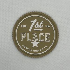 May include: A circular olive green rubberized patch with a serrated edge. The text reads 1st PLACE in white, with ESTD 1984 on the sides. A white star is centered below the text, and the words AWARDS AND GIFTS are printed in a white arc at the bottom.