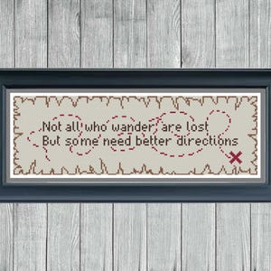 May include: Cross-stitch art in a dark blue frame, featuring the quote "Not all who wander are lost, but some need better directions." The design includes a map-like dotted line and a red "X" on a beige background, set against a wood-grain backdrop.