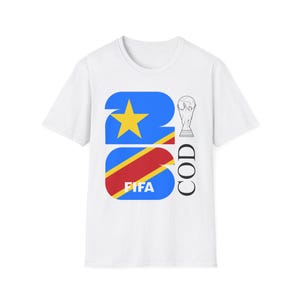 Puede incluir: Camiseta blanca con un diseño gráfico que incorpora la bandera de la República Democrática del Congo, una estrella dorada y el texto "FIFA". El diseño también incluye el texto "COD" y un trofeo.