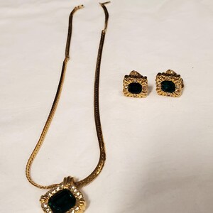 May include: A gold chain necklace with a square pendant featuring a large green gemstone surrounded by smaller clear gemstones. Matching earrings with square green gemstones.