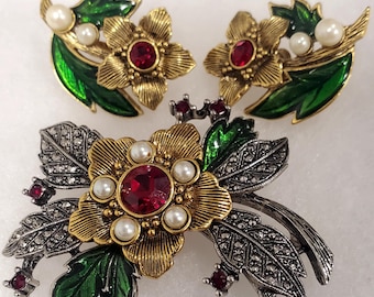 May include: A set of gold tone earrings and a brooch featuring a red rhinestone flower with green enamel leaves and pearl accents.