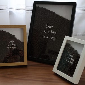 Könnte beinhalten: Eine weiße Keramik-Tasse mit schwarzem Henkel und dem Text "But First, Coffee" in handschriftlichem Stil. Die Tasse steht auf einer weißen Oberfläche. Die Tasse ist etwa 10 cm hoch.